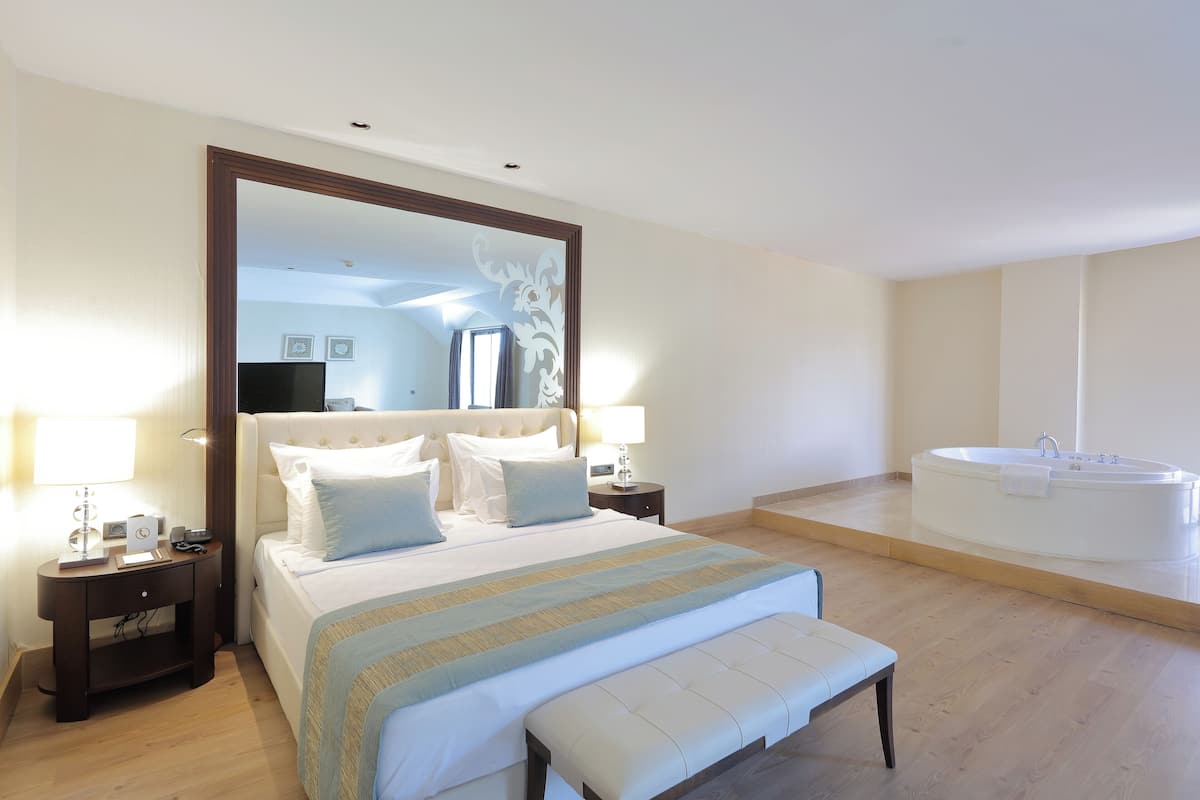 A spacious bedroom with a large bed, a bedside table, a nightstand, a bench, a large mirror, and a separate bathroom with a bathtub.