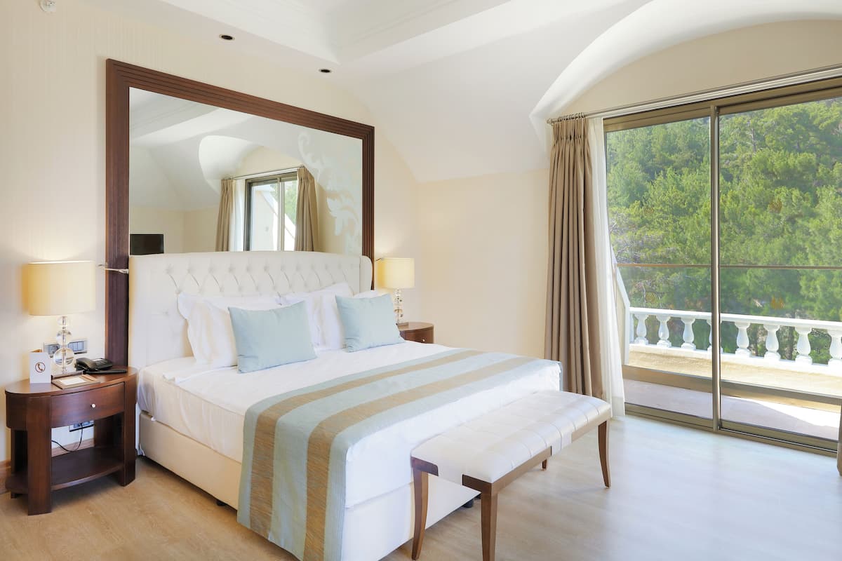 A spacious bedroom with a large bed, a nightstand, a bench, and a balcony with a view of trees.