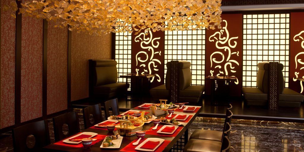 A traditional dining room with a long table set for a meal, featuring red and white tableware, and a decorative ceiling light fixture.
