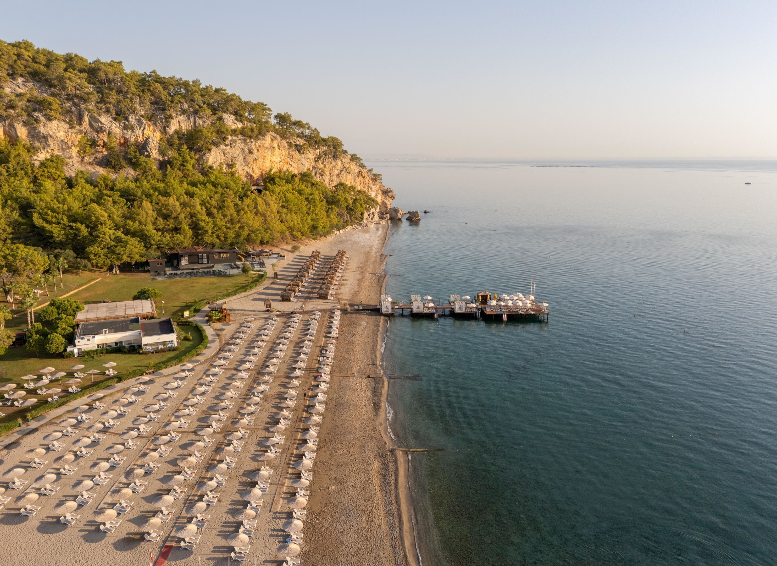 A coastal hotel with rows of beachside rooms, a pier extending into the sea, and a mountainous backdrop.