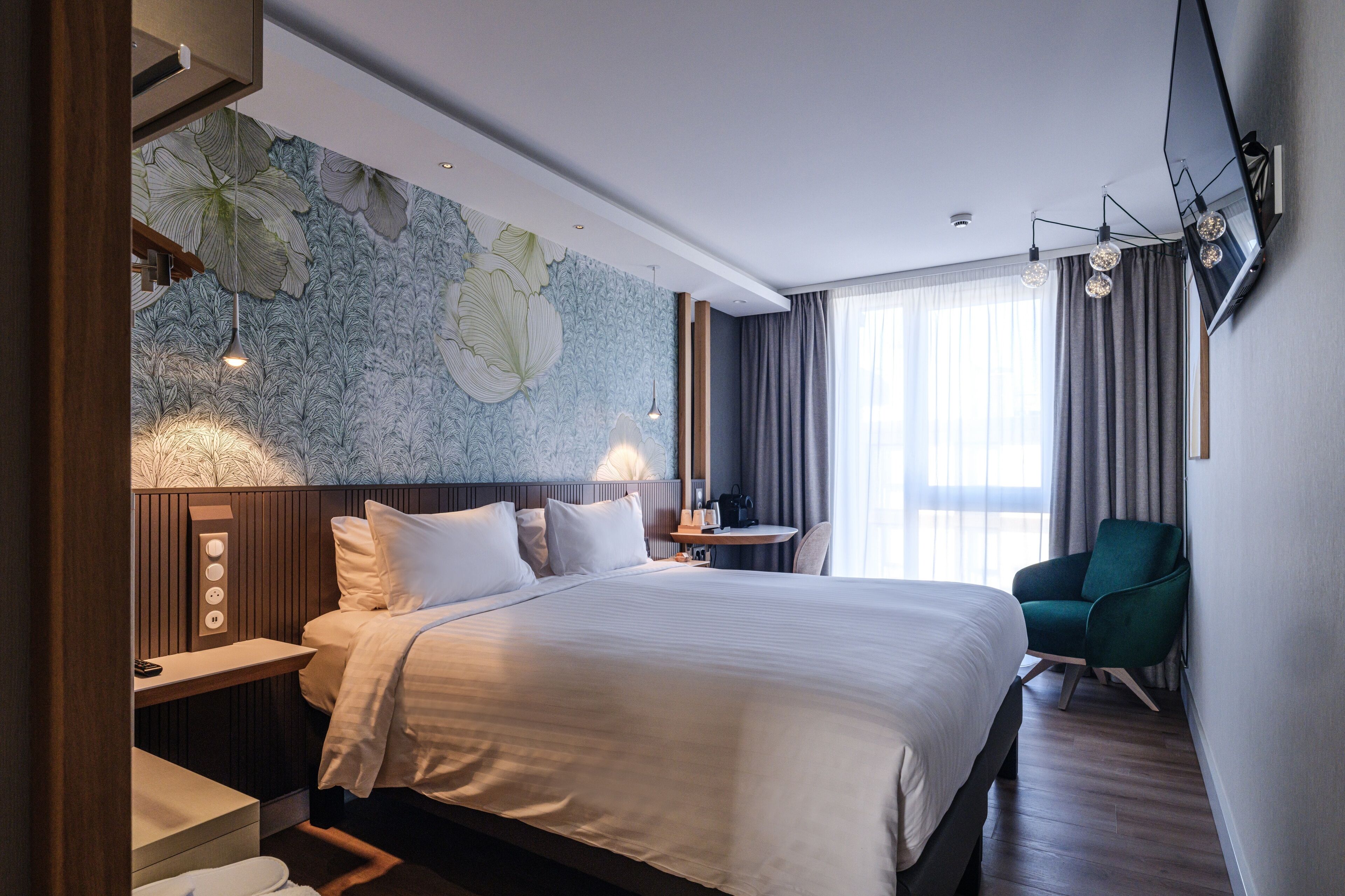 A hotel room with a large bed, a desk, a chair, a small table, and a wall with a floral wallpaper.