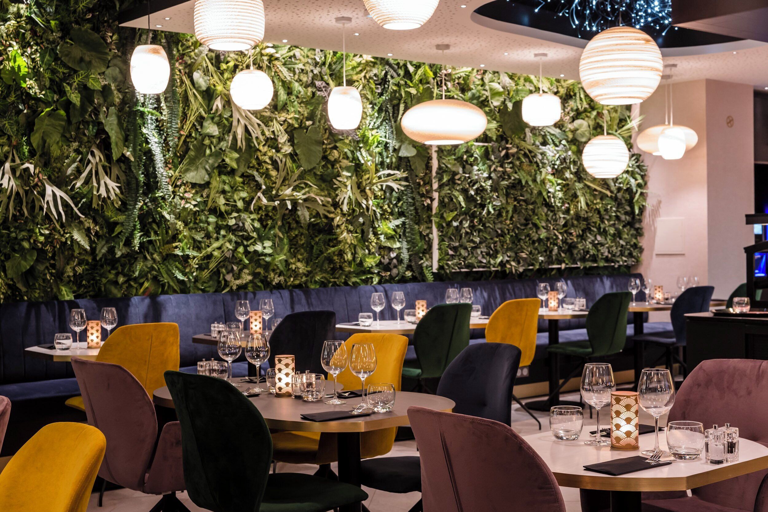 Een restaurantinterieur met ronde tafels, stoelen in verschillende kleuren en een wand vol begroeiing.