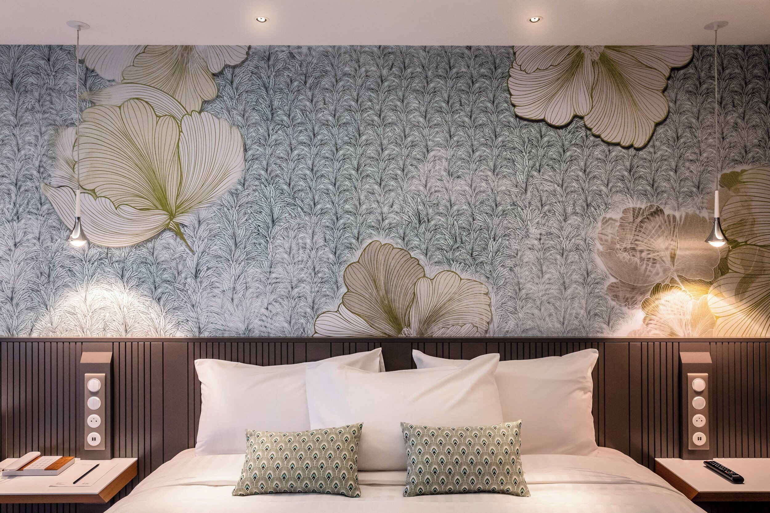 A bedroom with a large floral wallpaper, a bed with white linens, two pillows, bedside tables with lamps, and a wall-mounted power outlet.