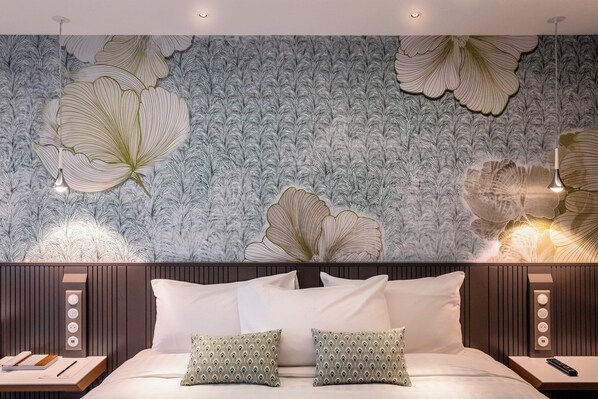 A bedroom with a large floral wallpaper, a bed with white linens, two pillows, bedside tables with lamps, and a wall-mounted power outlet.