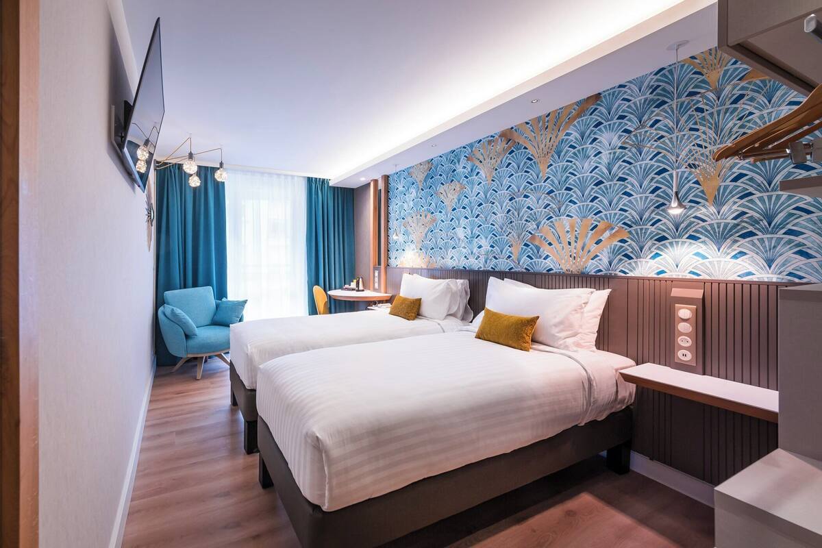 A modern hotel room with two beds, a seating area with a chair, a table, and a decorative wall.