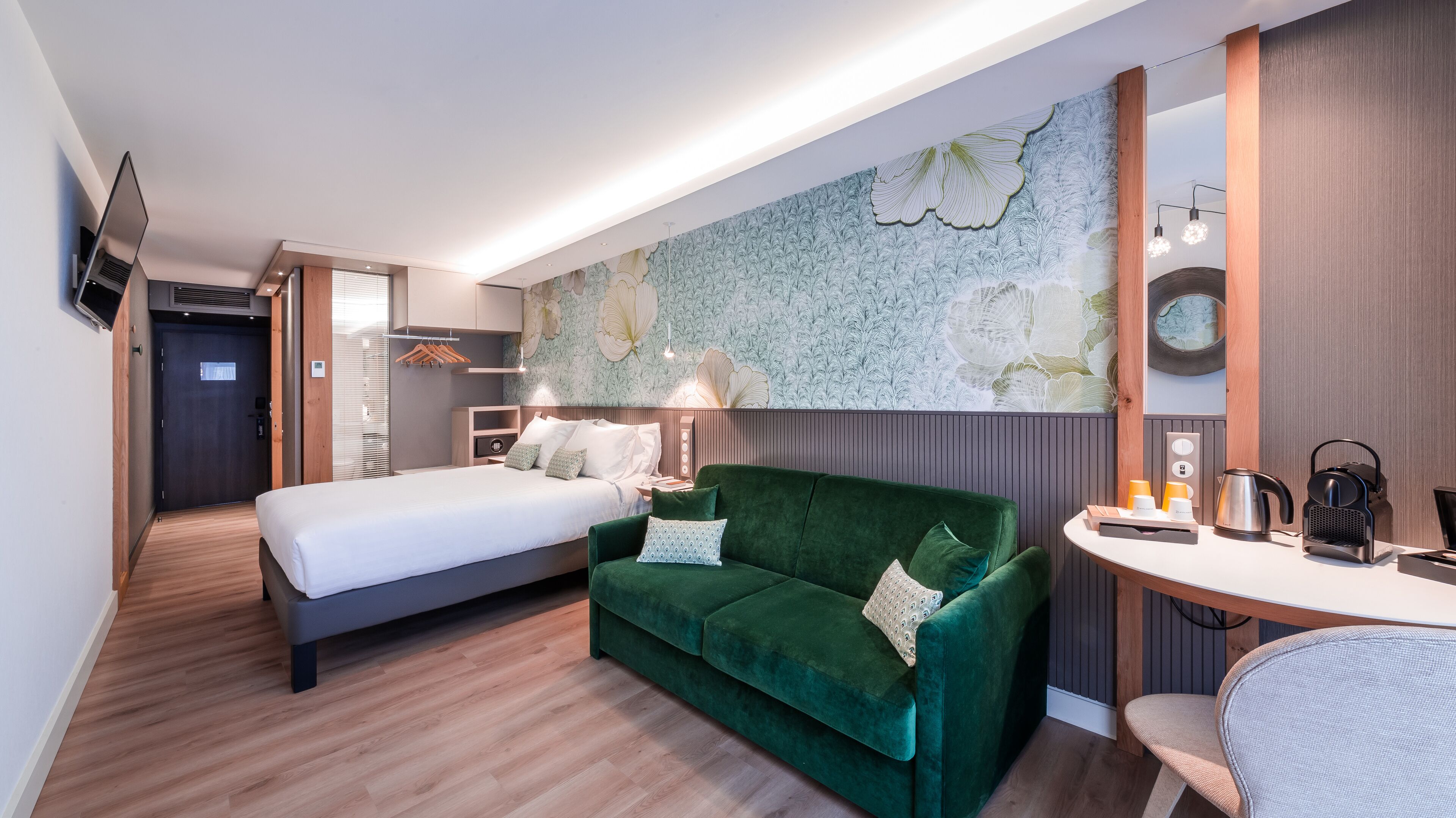 Une chambre d’hôtel moderne avec un lit, un canapé vert, un bureau avec une bouilloire et un grand papier peint à motifs floraux.