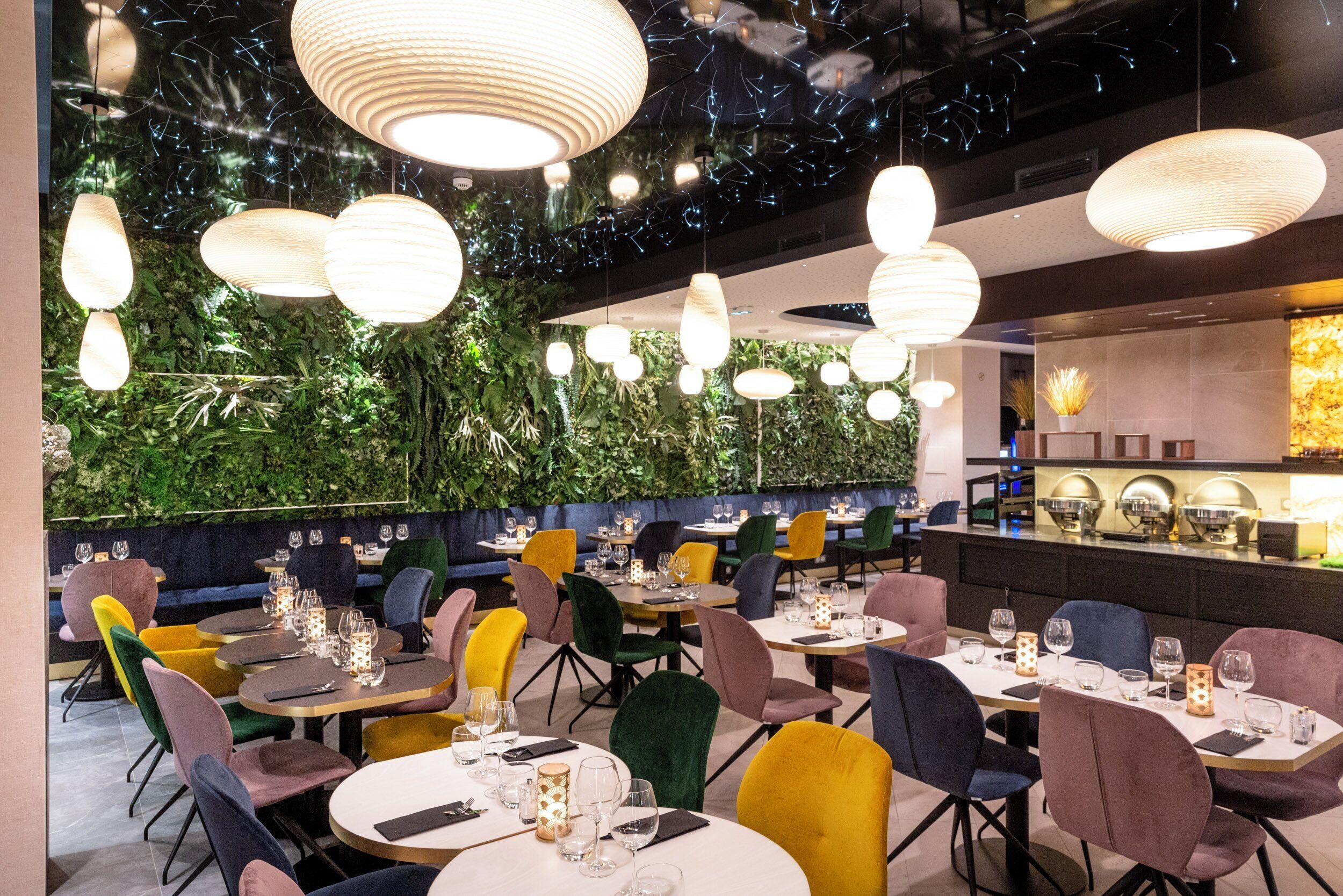 Ein Restaurantinnenraum mit runden Tischen, bunten Stühlen, hängenden Pendelleuchten und einer grünen Wand mit Pflanzen.