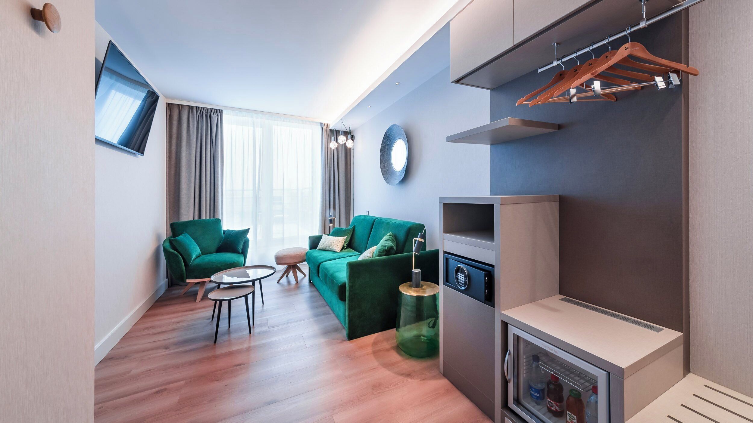 Ein modernes Wohnzimmer mit einem grünen Sofagarnitur, einem kleinen runden Tisch, einem flachbildfernseher an der Wand und einem eingebauten Minikühlschrank.