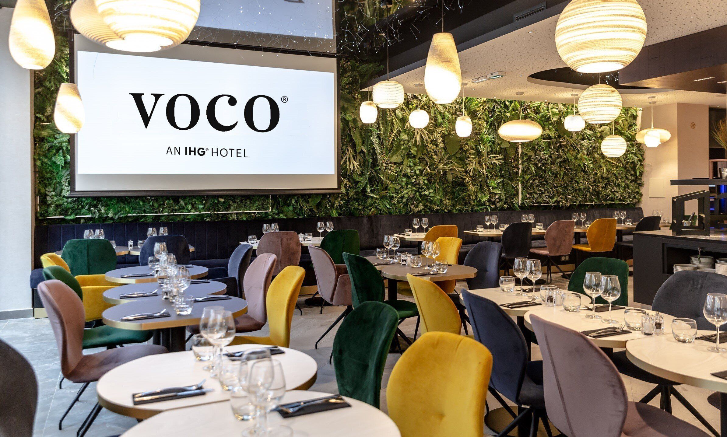 A restaurant area with tables set for dining, a large screen displaying 'VOCO AN IHG HOTEL', and hanging pendant lights.