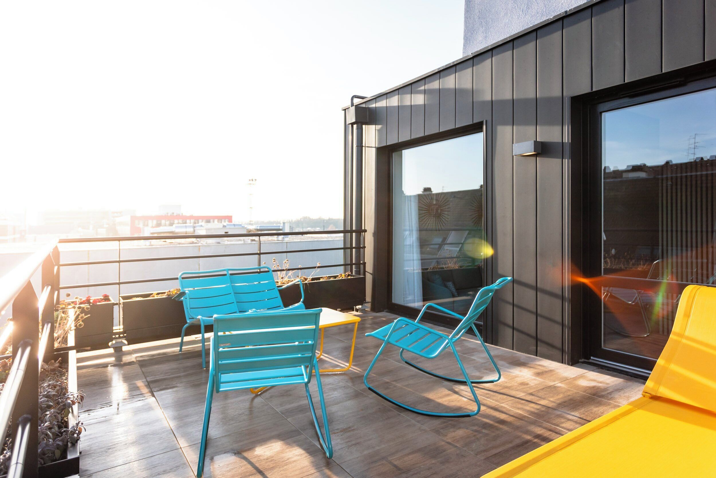A rooftop terrace with two blue rocking chairs and a yellow sofa, overlooking a cityscape.