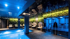 Een modern binnenzwembad met loungebedden, een glazen wand en een plafond met hangende lampen.