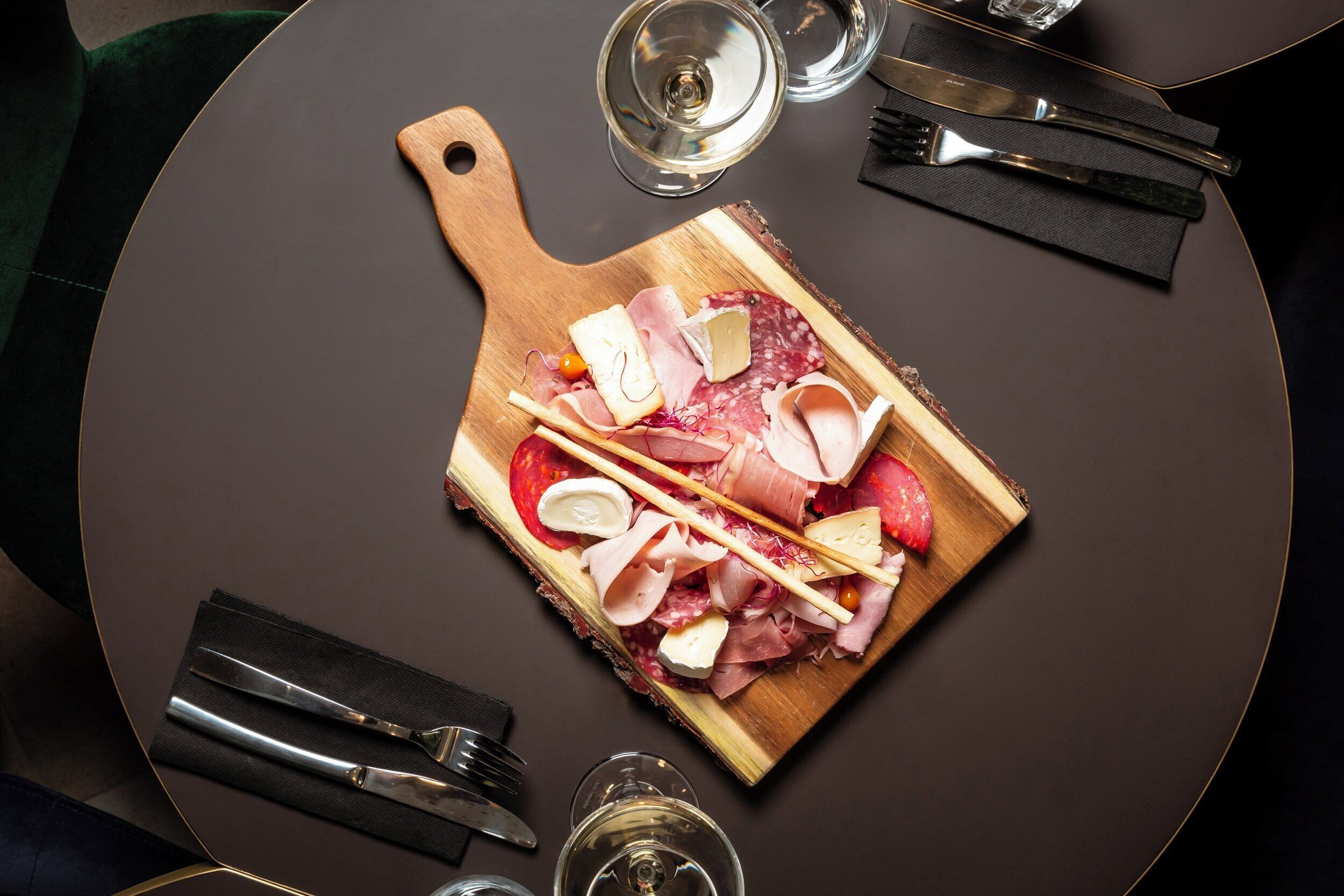 Una mesa redonda con una tabla de madera que sostiene varios tipos de carne y queso, acompañada de cubiertos y vasos.