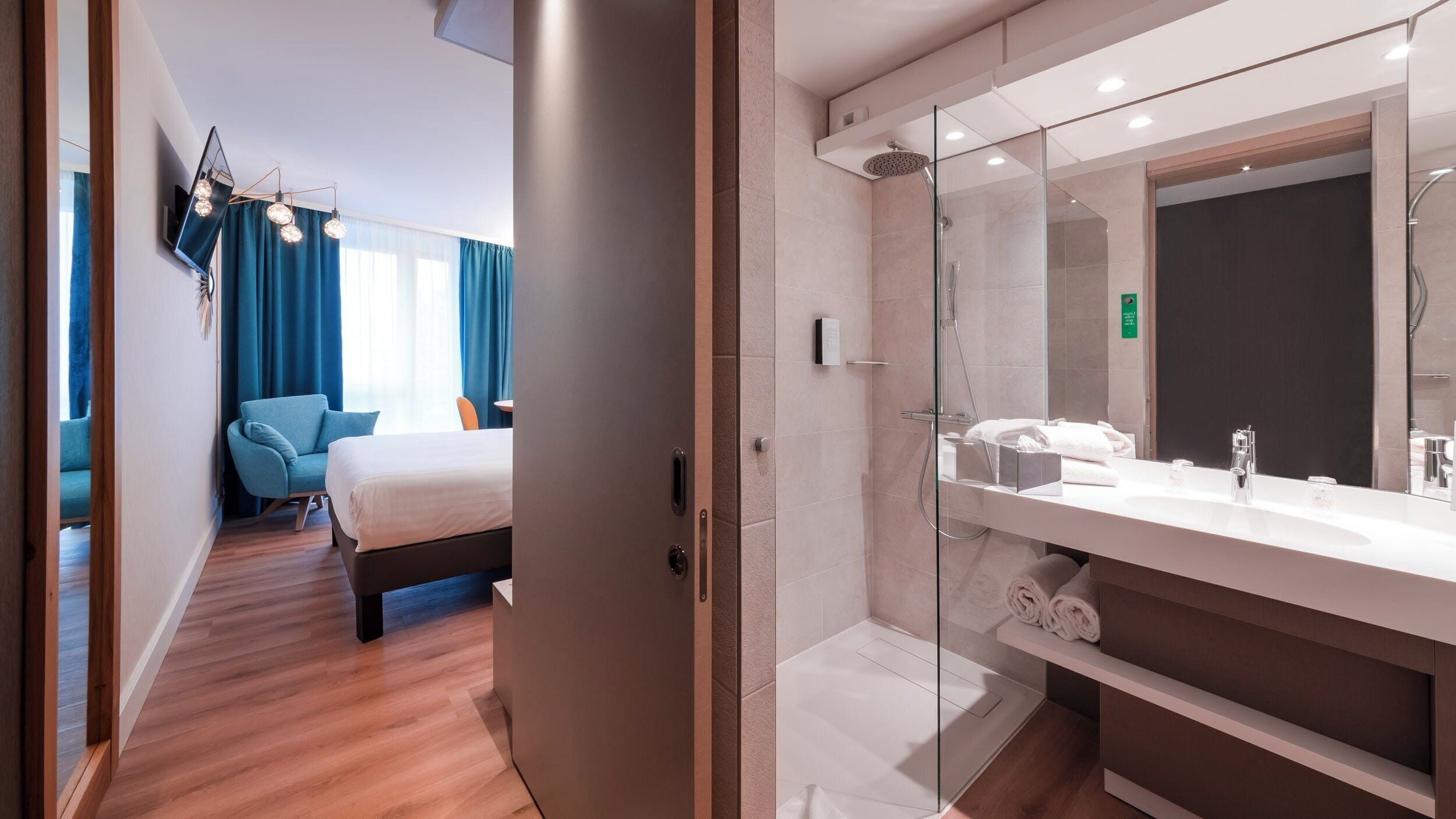 A hotel room with a bed, a seating area with a chair, a large bathroom with a glass shower enclosure, and a modern design.