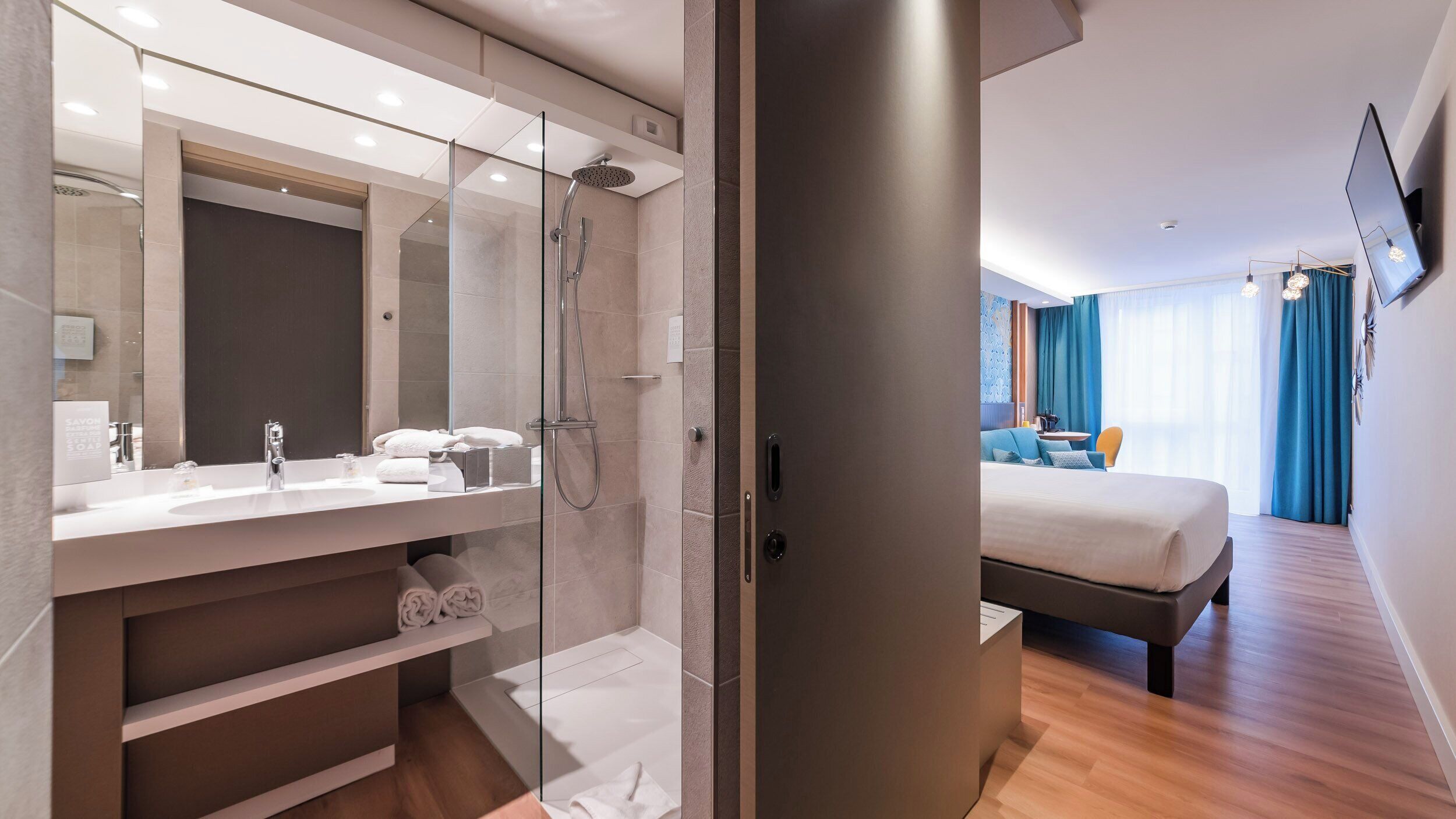A modern hotel room with a glass-enclosed shower, a large bed, a bedside table, and a TV mounted on the wall.