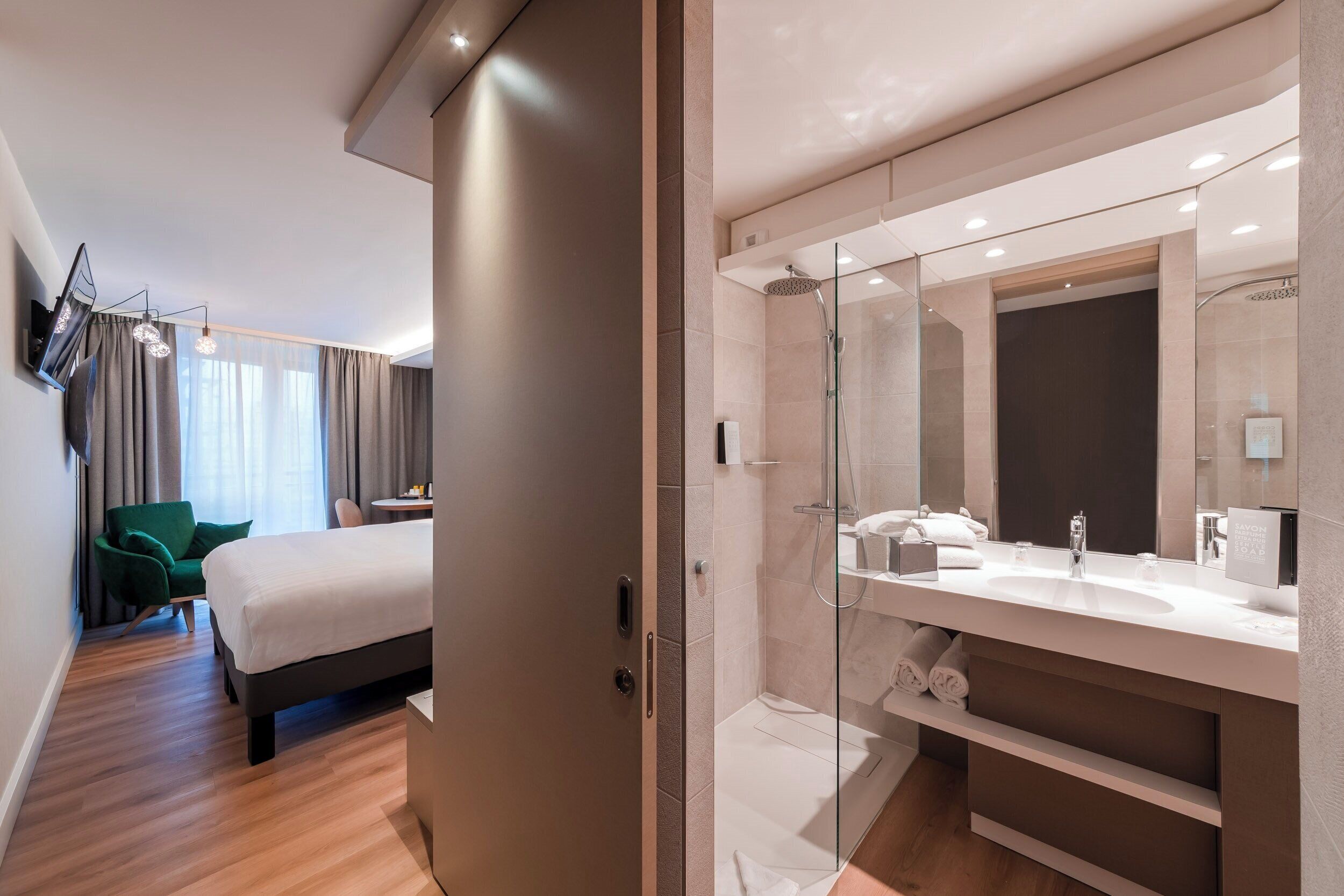 A modern hotel room with a large bed, a separate bathroom with a glass shower enclosure, and a green armchair.
