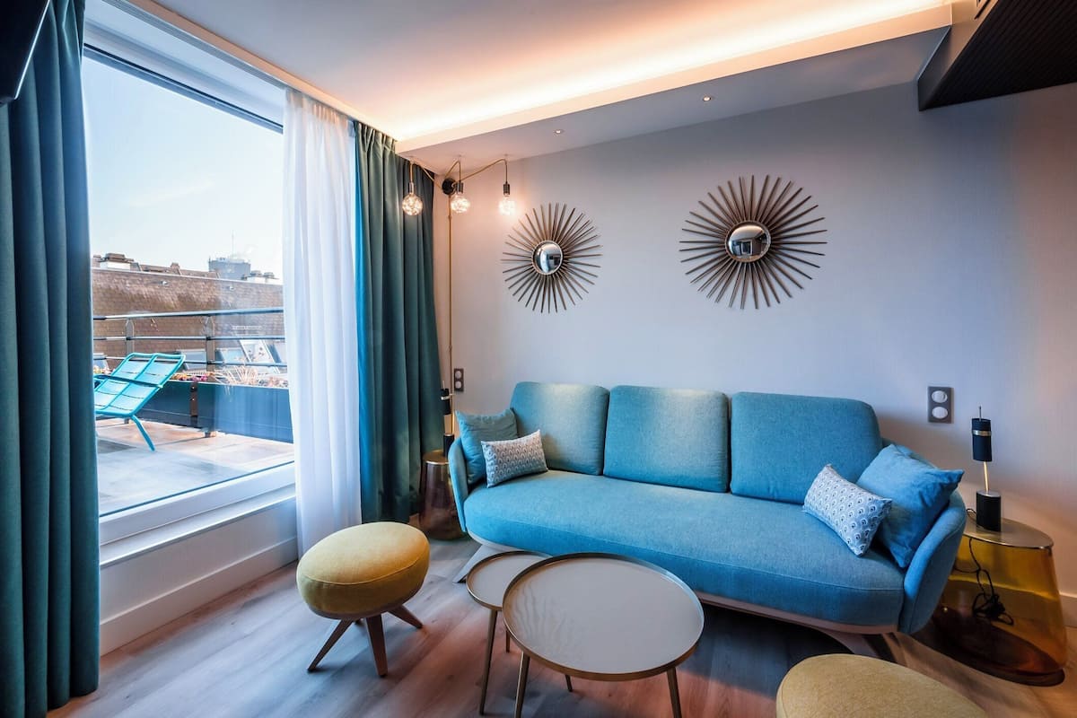 A modern living room with a blue sofa, two small round tables, and a yellow ottoman. There are two wall-mounted decorative mirrors and a floor lamp.