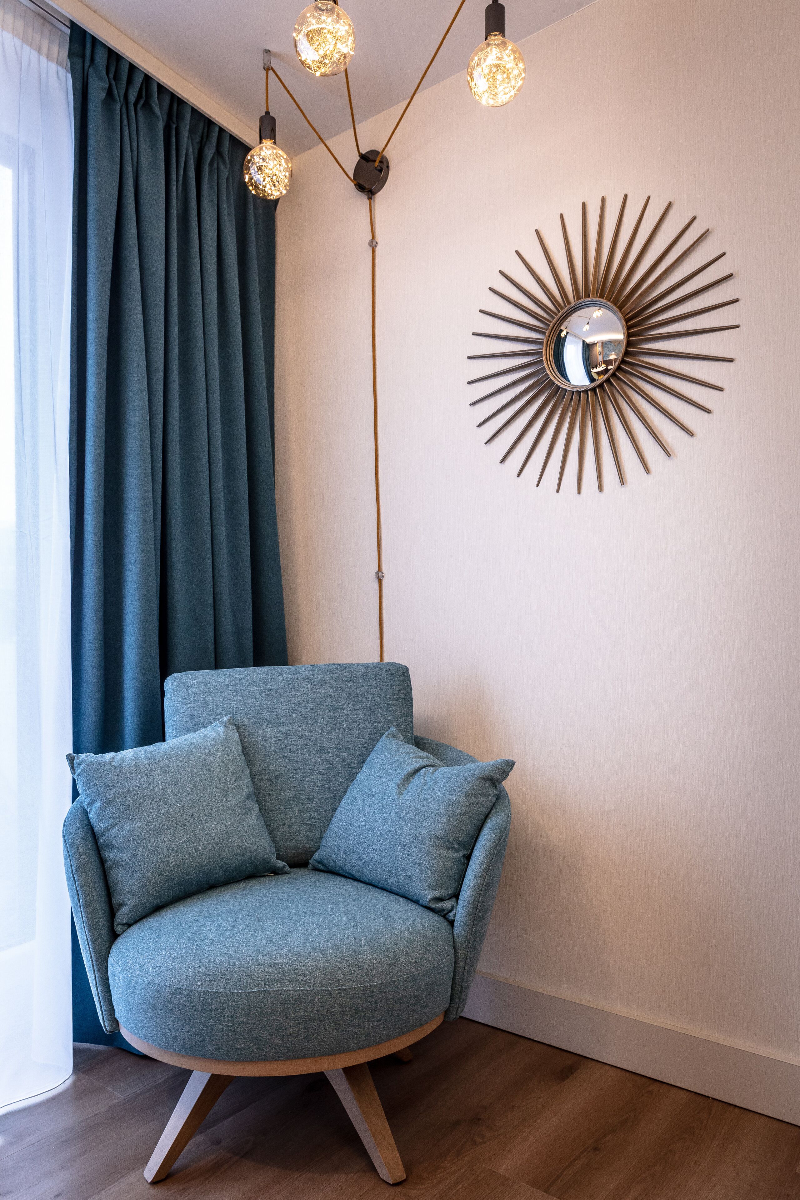 Ein modernes Wohnzimmer mit einem grauen Sessel, einem Spiegel im Sonnenstrahlendesign und einer hängenden Leuchte.