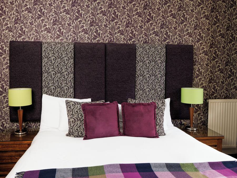 A neatly made bed with a white duvet, two purple pillows, and a colorful blanket. There are two bedside tables with lamps, and the walls have a patterned wallpaper.