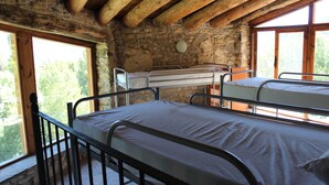 A rustic cabin with two metal bunk beds, stone walls, and large windows offering a view of greenery. - Albergue Casa Fumenal (Valle de Lierp)