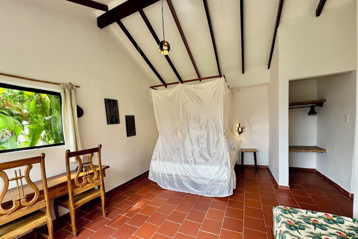 A room with a bed under a white canopy, a wooden bench, a small table, and a view of greenery through a window.