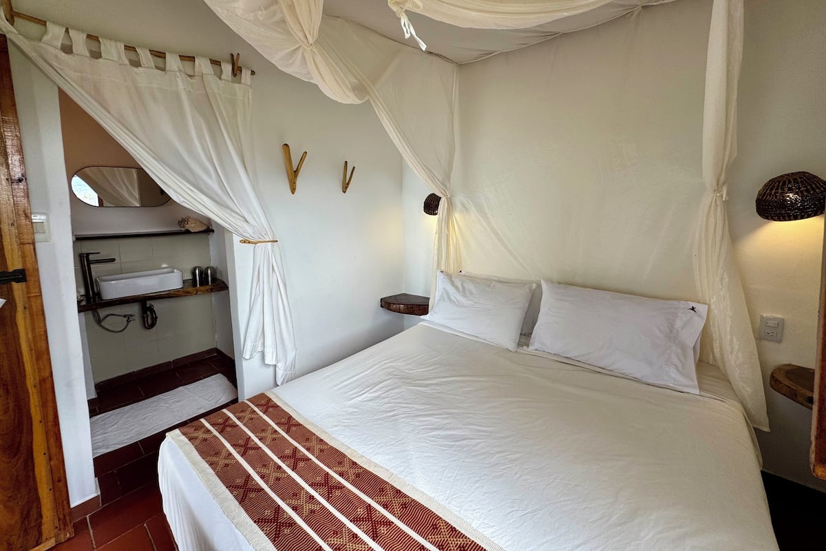 A bedroom with a bed, white curtains, a bedside table, a mirror, and a small shelf with toiletries.