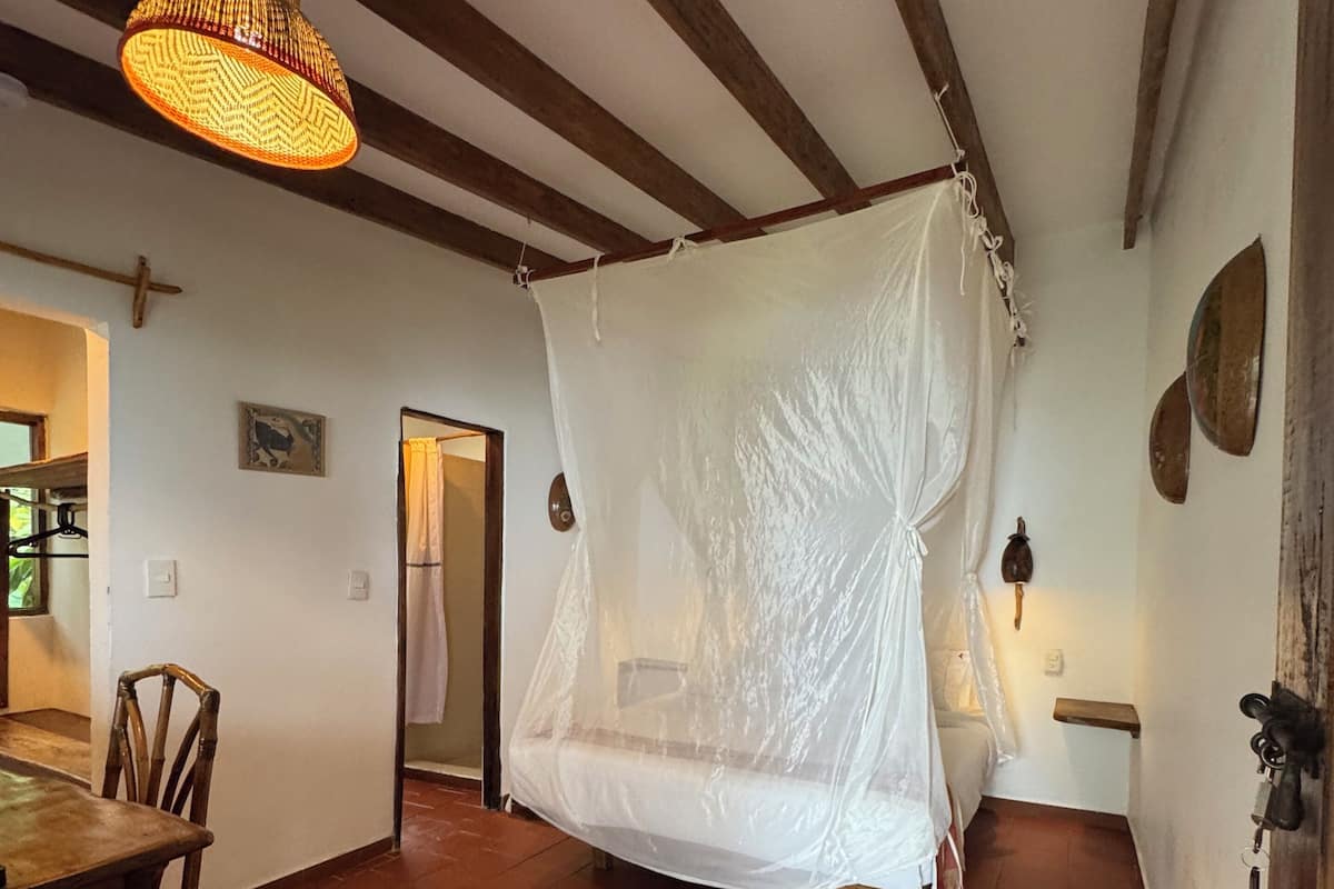 A rustic room with a bed covered in a white canopy, wooden beams on the ceiling, and a woven light fixture.