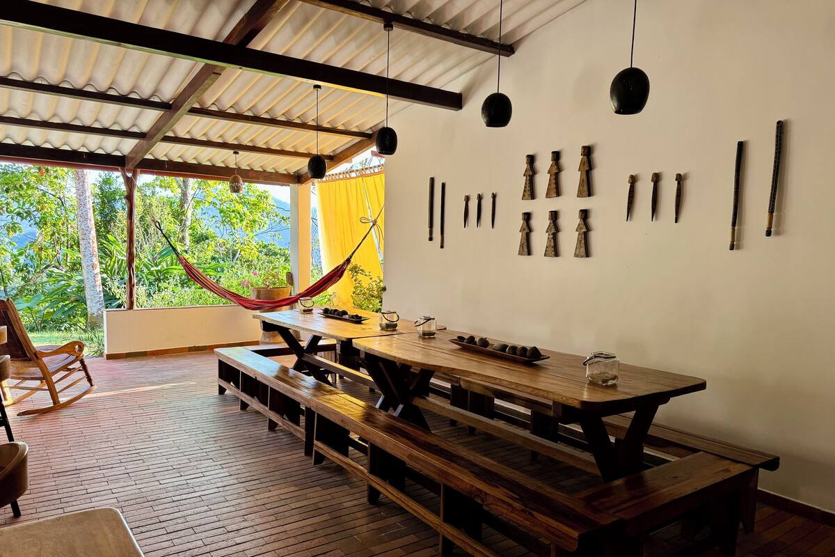 A wooden dining table with benches, a hammock, and a view of greenery through large windows.