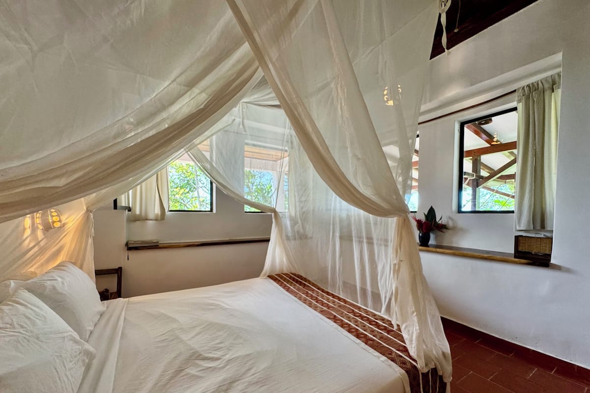 A bedroom with a bed under a canopy, a window with curtains, and a brick wall.