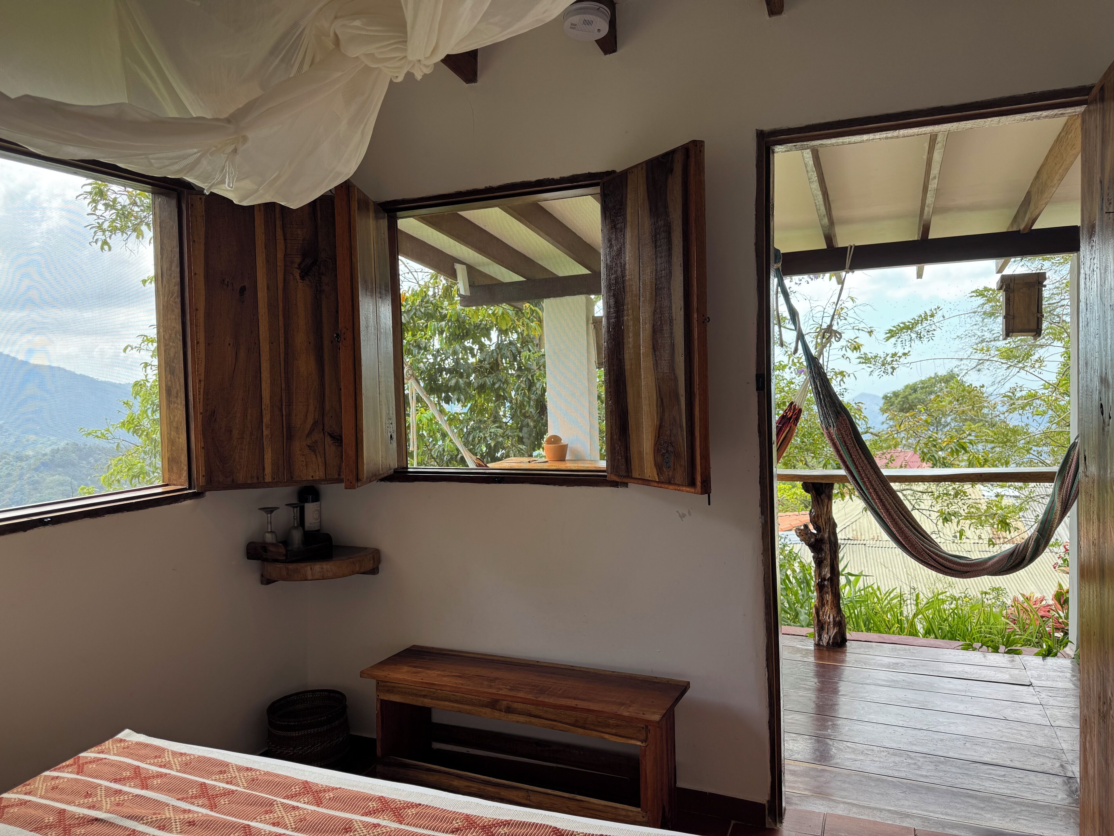 A bedroom with a large window, wooden furniture, and a hammock outside.