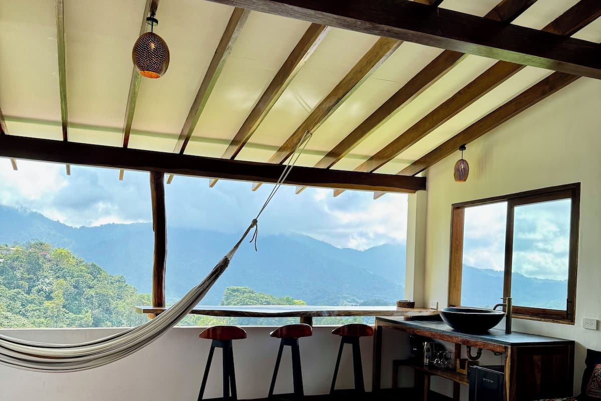 A room with a hammock, a wooden table, a sofa with a floral pattern, a kitchen area with a stove, and a view of mountains.