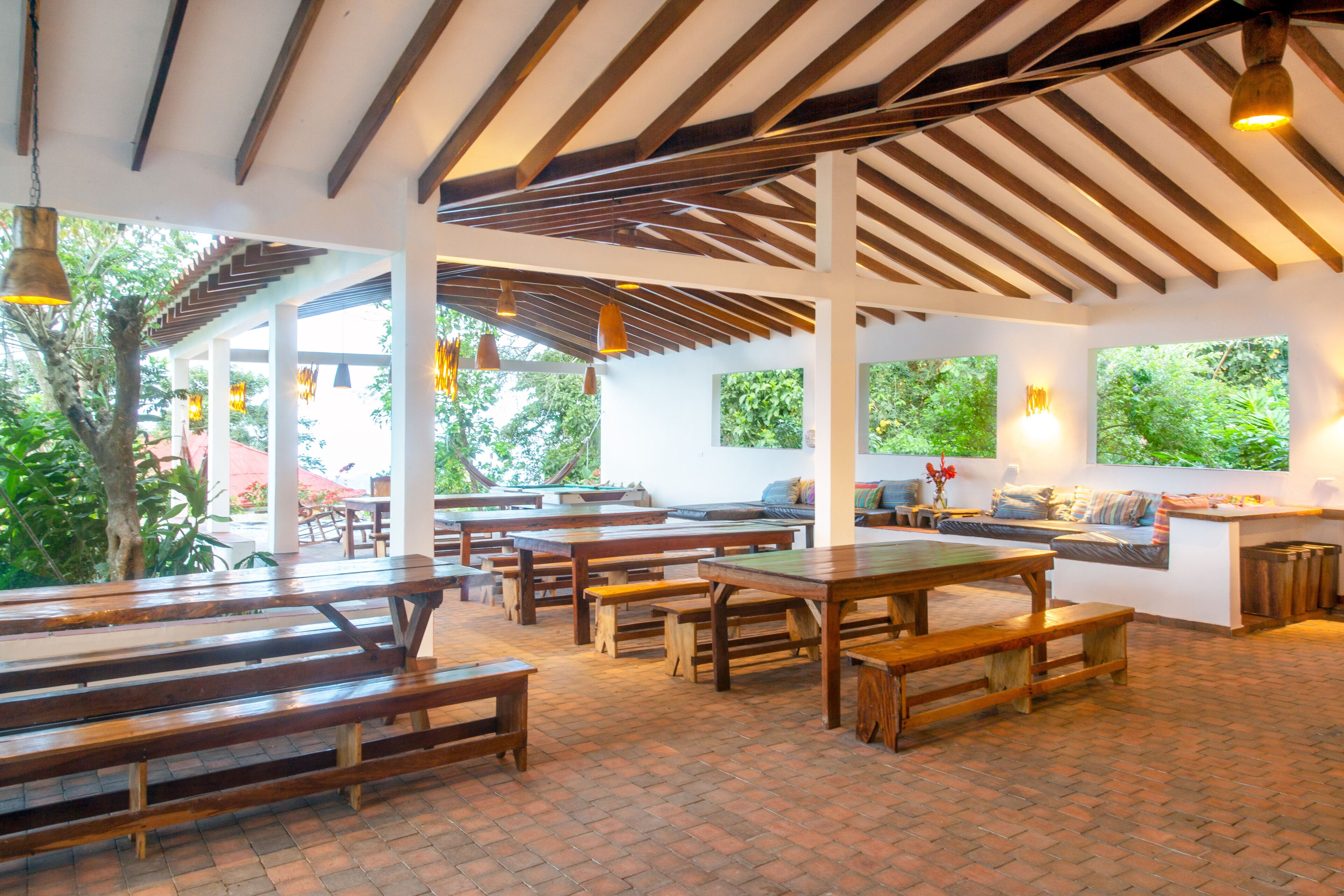 An indoor dining area with wooden benches and tables, large windows, and a brick floor.