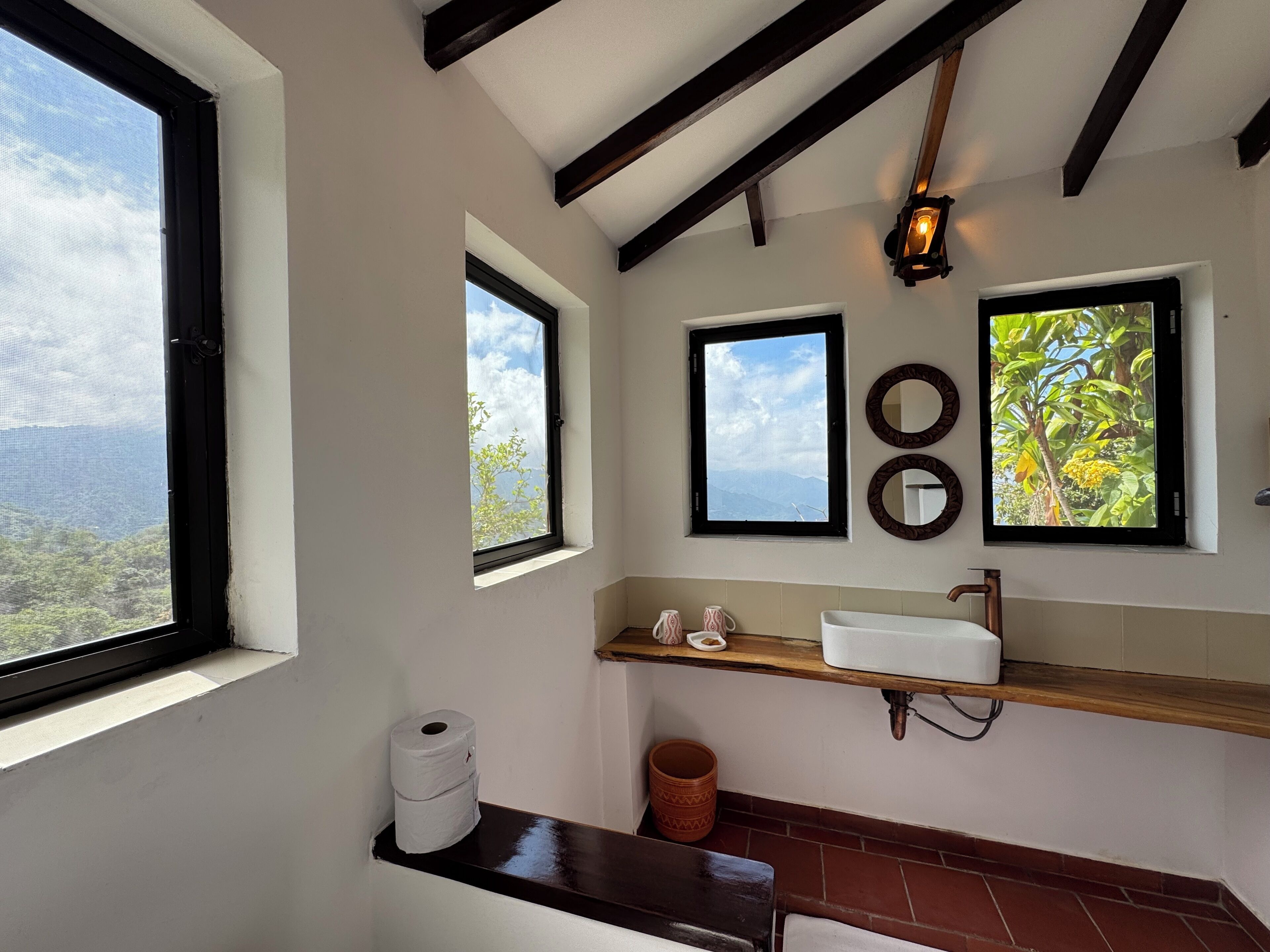 A bathroom with a window, a shelf with toiletries, a mirror, and a hanging light fixture.