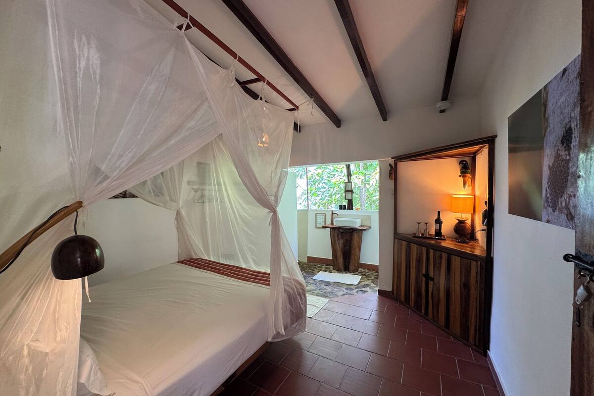 A bedroom with a four-poster bed, a small bathroom with a sink and mirror, and a view of greenery outside.