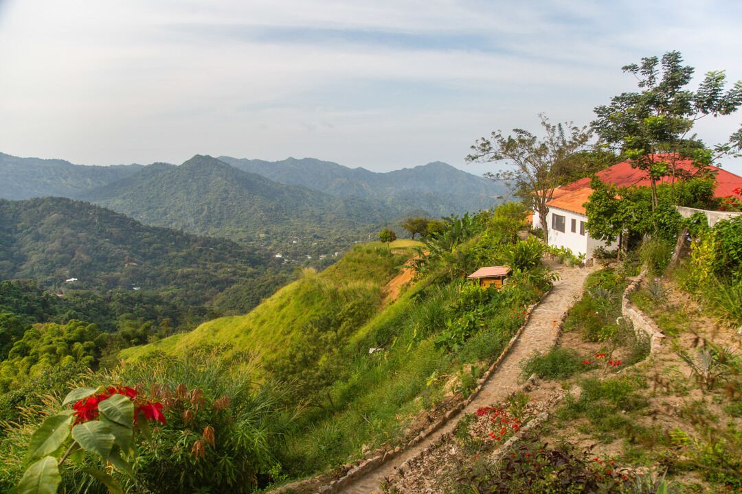 Une maison située sur une colline, avec un toit rouge, entourée d’une végétation luxuriante et avec des montagnes en arrière-plan.