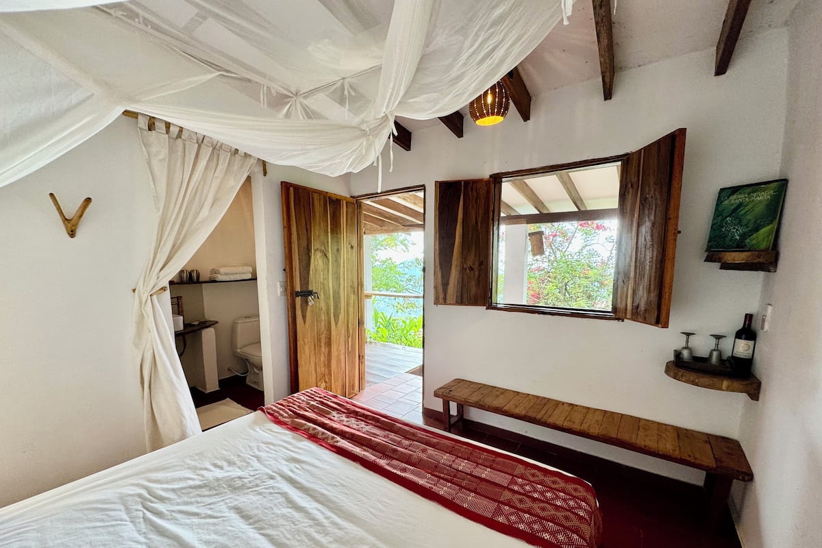 A bedroom with a bed, a bench, a window, a mirror, and a shelf with bottles.