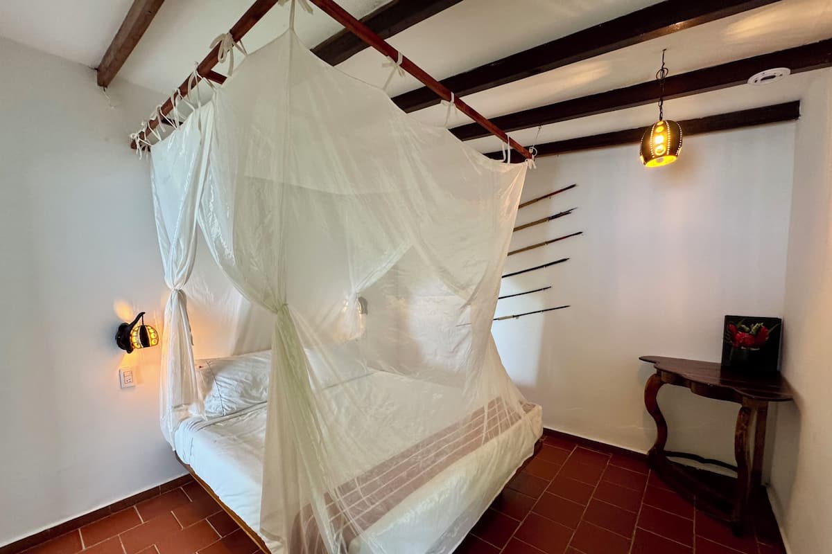 A room with a bed under a white canopy, a wall-mounted light, and a small table with a picture.