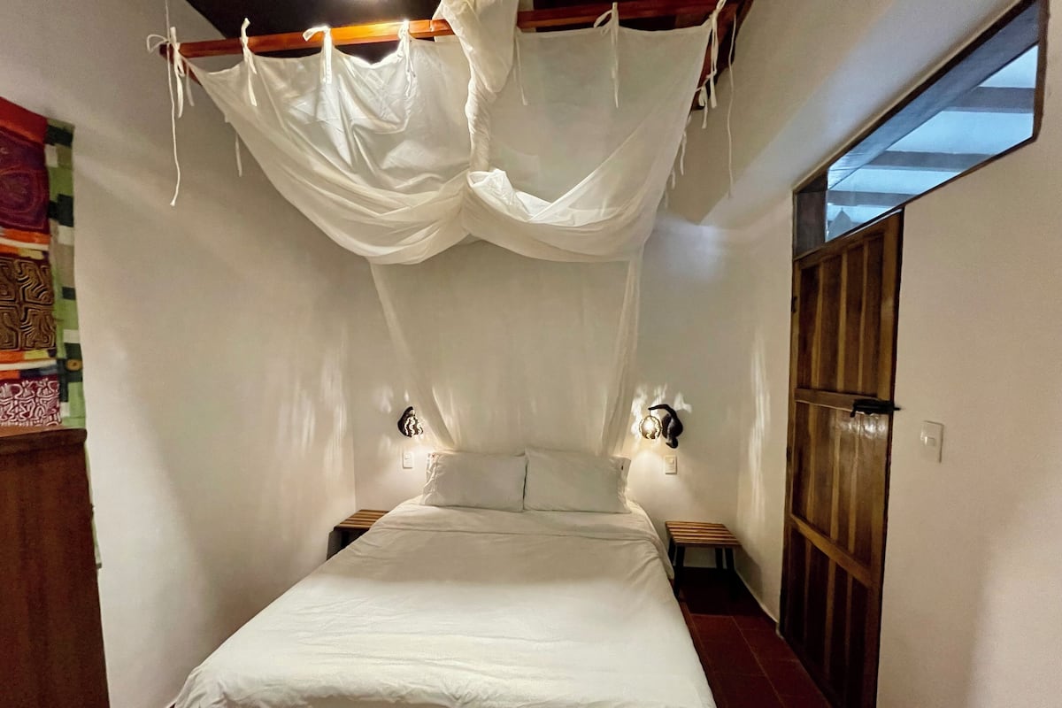 A bedroom with a bed, white curtains, two bedside lamps, a wooden headboard, and a window.