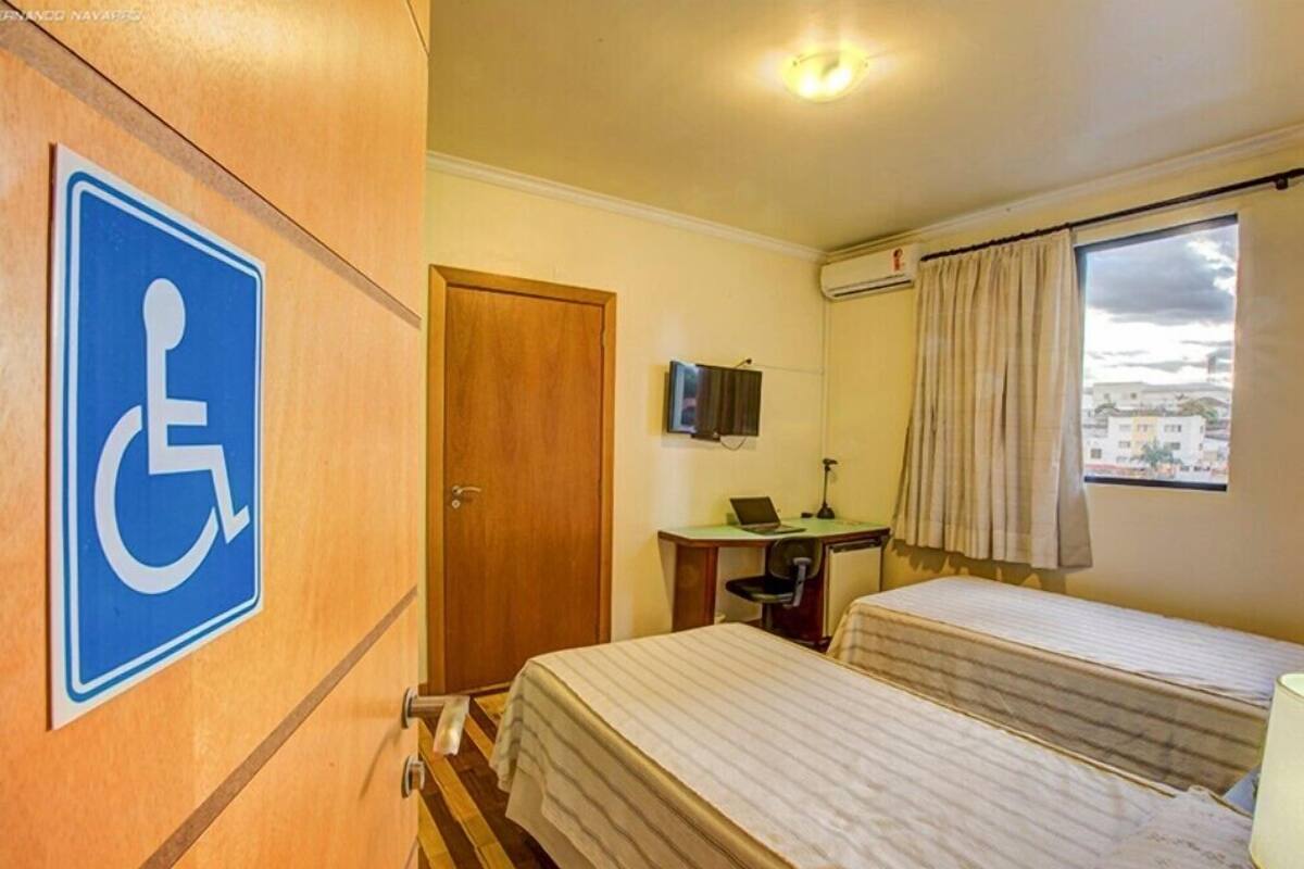 Ein Hotelzimmer mit Rollstuhlgerechterkeitssymbol, einem Schreibtisch mit einem Laptop, zwei Betten und einem Fenster mit Vorhängen.