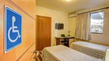 Quarto de hotel com sinal de acessibilidade para cadeiras de rodas, uma mesa com um laptop, duas camas e uma janela com cortinas.