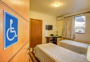 A hotel room with a wheelchair accessibility sign, a desk with a laptop, two beds, and a window with curtains. - Almasty Hotel (Chapecó)