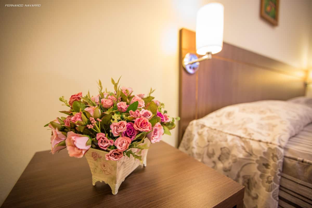 Ein Strauß rosa Rosen in einer Zierdekor-Vase auf einem Holztisch neben einem Bett mit Kopfteil und einer Wandleuchte.