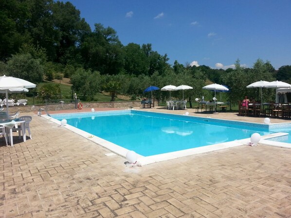 A rectangular swimming pool with a tiled deck, surrounded by white umbrellas and outdoor furniture.