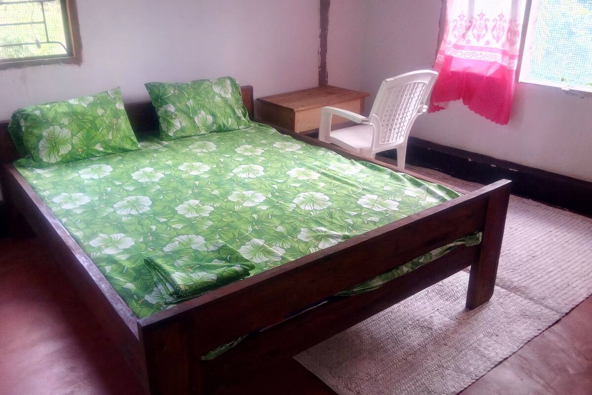 A room with a wooden bed, green floral bedspread, a white plastic chair, a small wooden table, and a window with a red curtain.