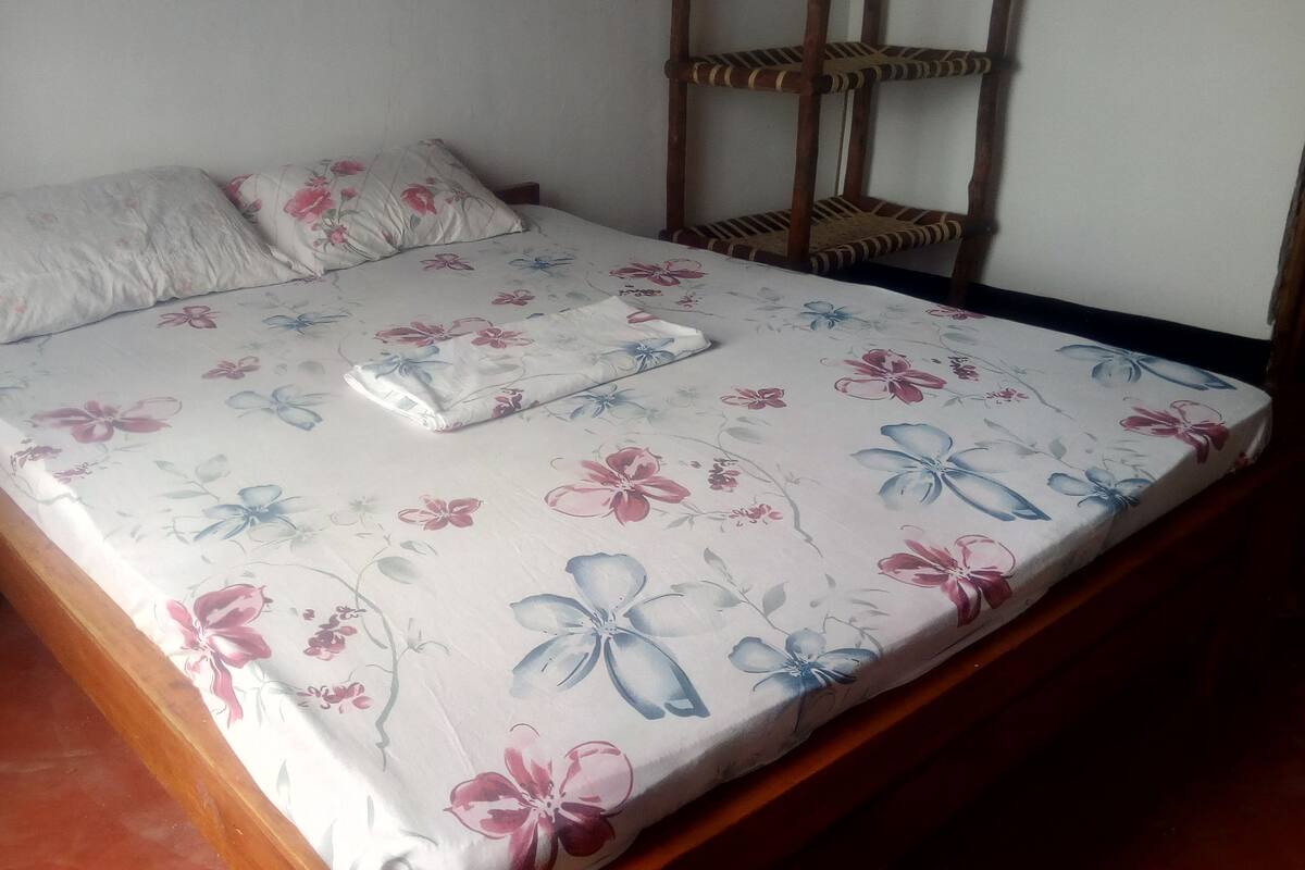 A bed with a floral bedspread and two pillows, a wooden nightstand, and a three-tiered wooden shelf.