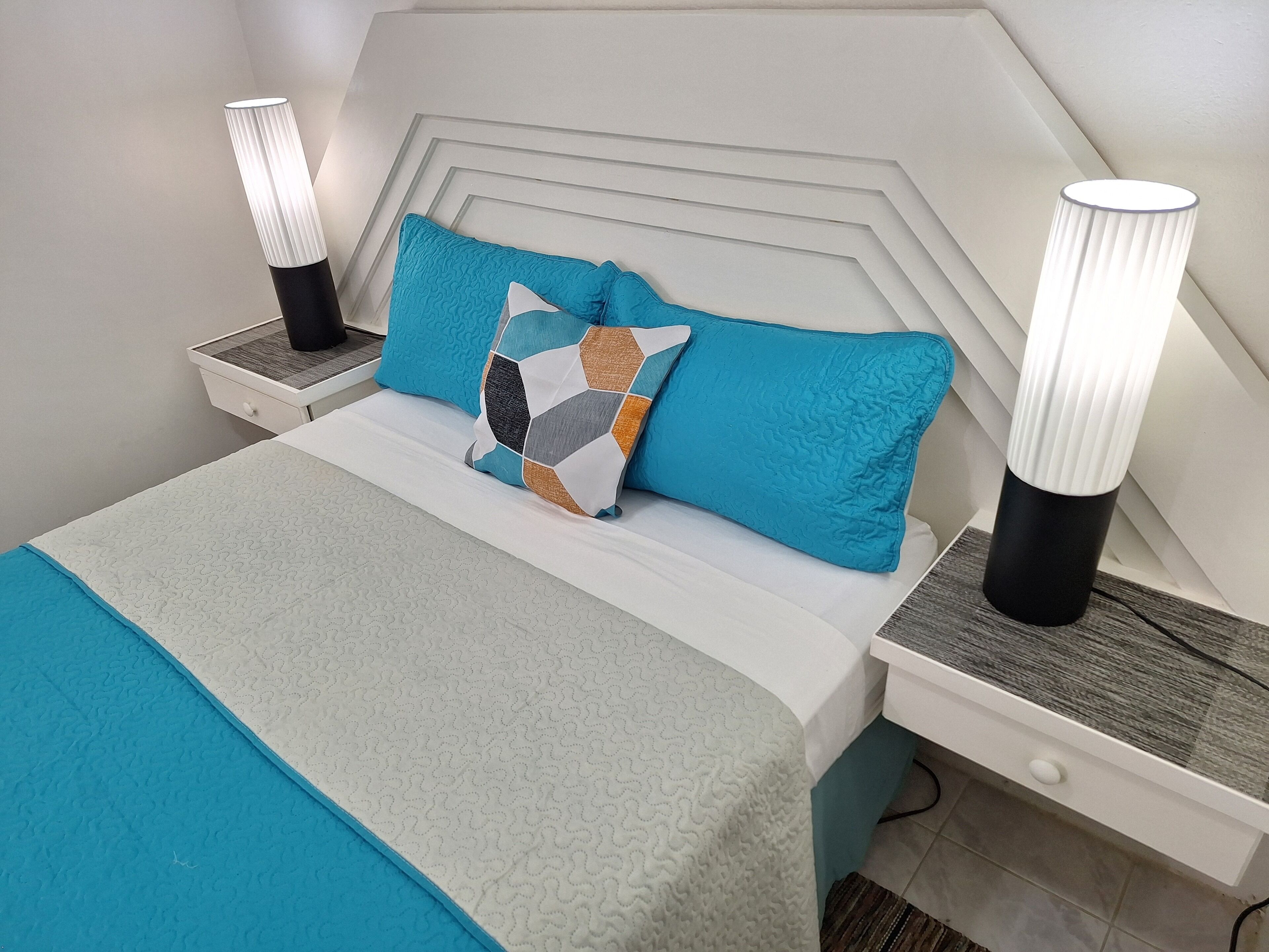 Un lit bien fait, recouvert d’une couvre-lit à motifs, avec un coussin aux formes géométriques et deux lampes de chevet posées sur des tables de chevet blanches.