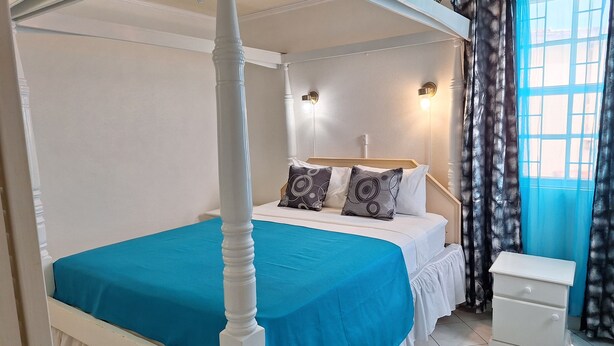 A four-poster bed with a blue bedspread, a bedside table, and a nightstand. - Cumber's Tropical Apartments (Oistins)
