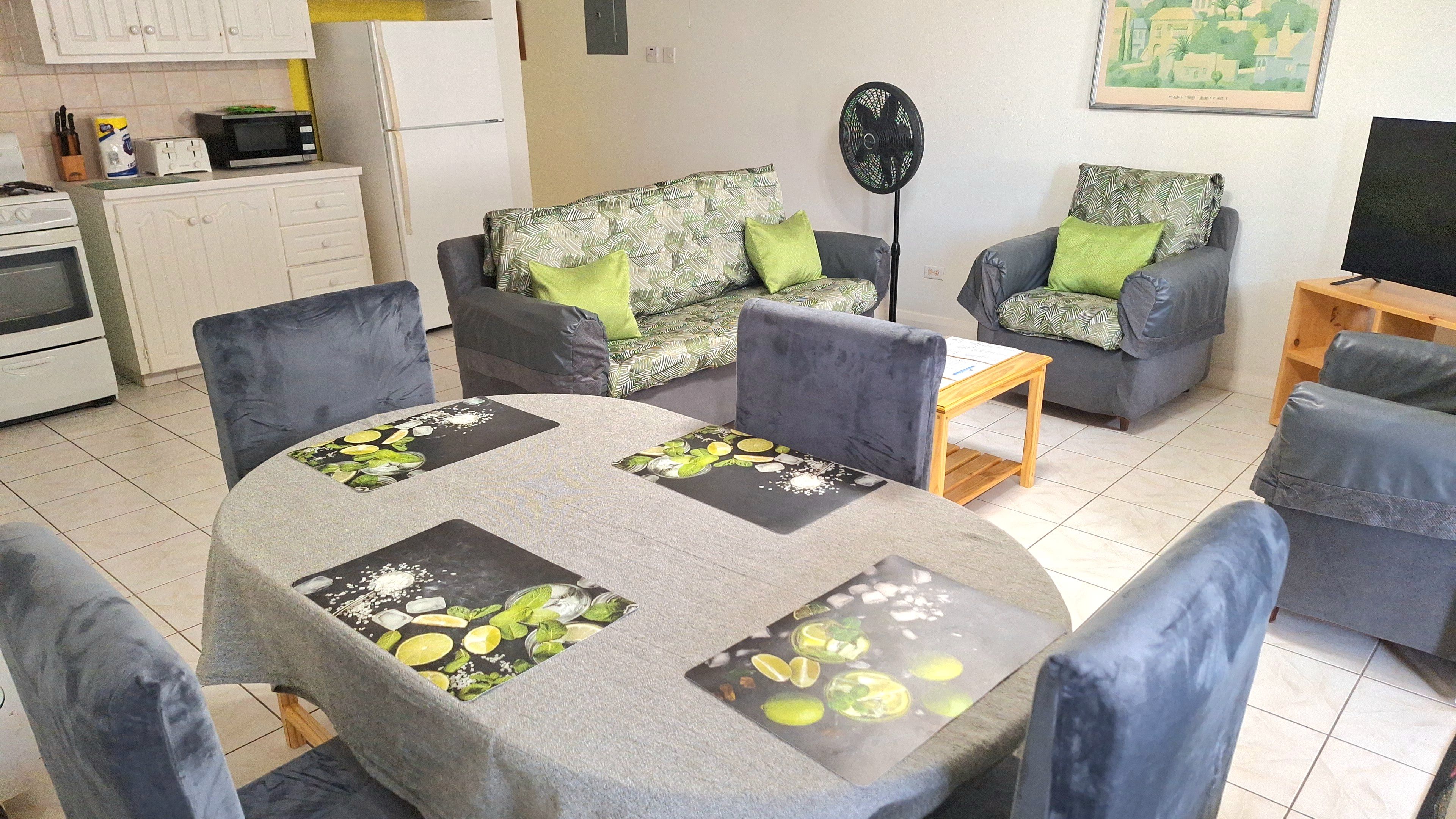 A living room with a dining table, two sofas with patterned cushions, a fan, and a kitchen area with appliances.
