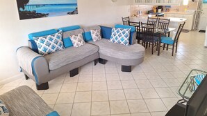A living room with a sectional sofa, dining table, chairs, ceiling fans, and a kitchen area in the background. - Cumber's Tropical Apartments (Oistins)