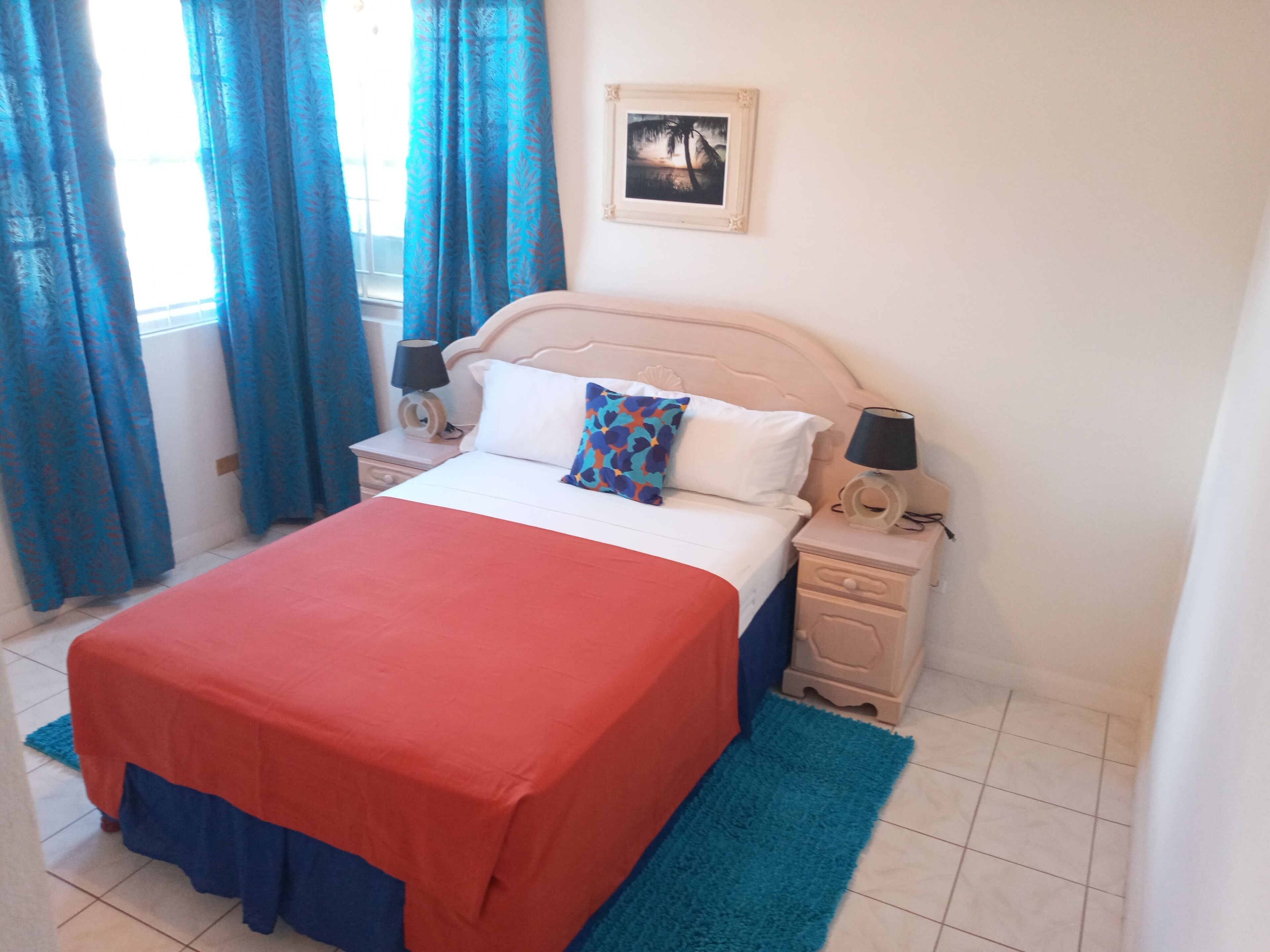 Une chambre à coucher avec un lit, deux tables de chevet, une commode, un tableau encadré au mur et des rideaux bleus.
