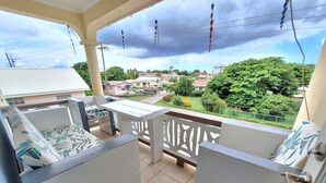 A balcony with a view of a residential area, featuring a white railing, a bench, and a door. - Cumber's Tropical Apartments (Oistins)